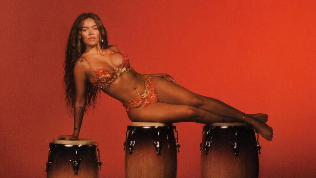 Karol G in a colorful bikini poses on three conga drums against a red background.