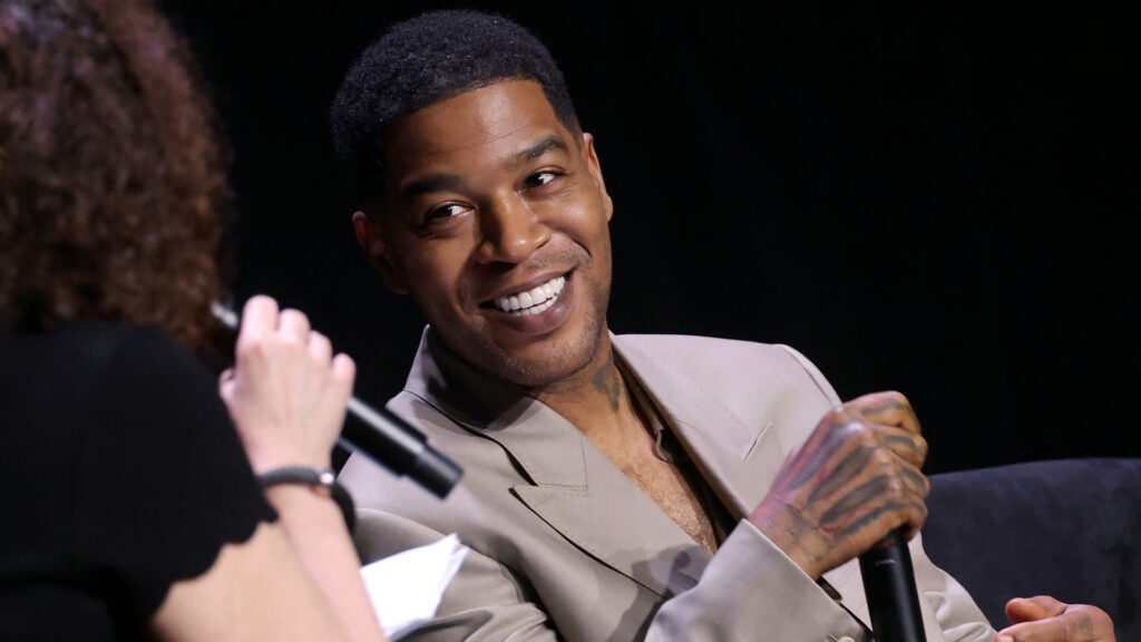 Kid Cudi, wearing a beige suit, smiles during an interview. He's holding a microphone, engaged in conversation with a woman.