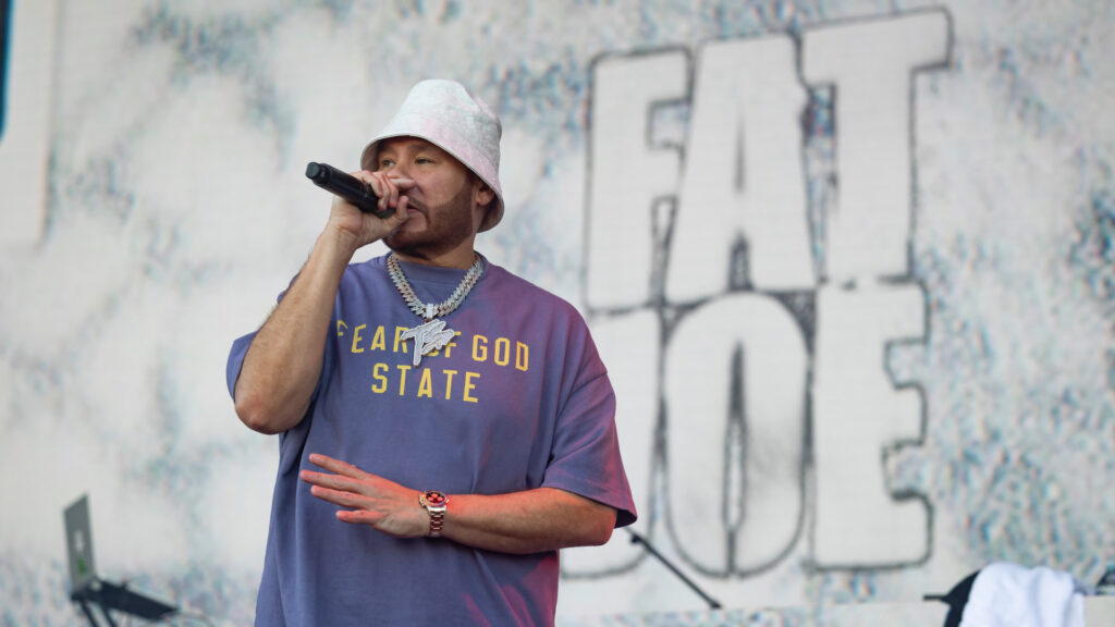 Fat Joe performing on stage, wearing a "Fear of God State" shirt and bucket hat, with his name displayed in the background.