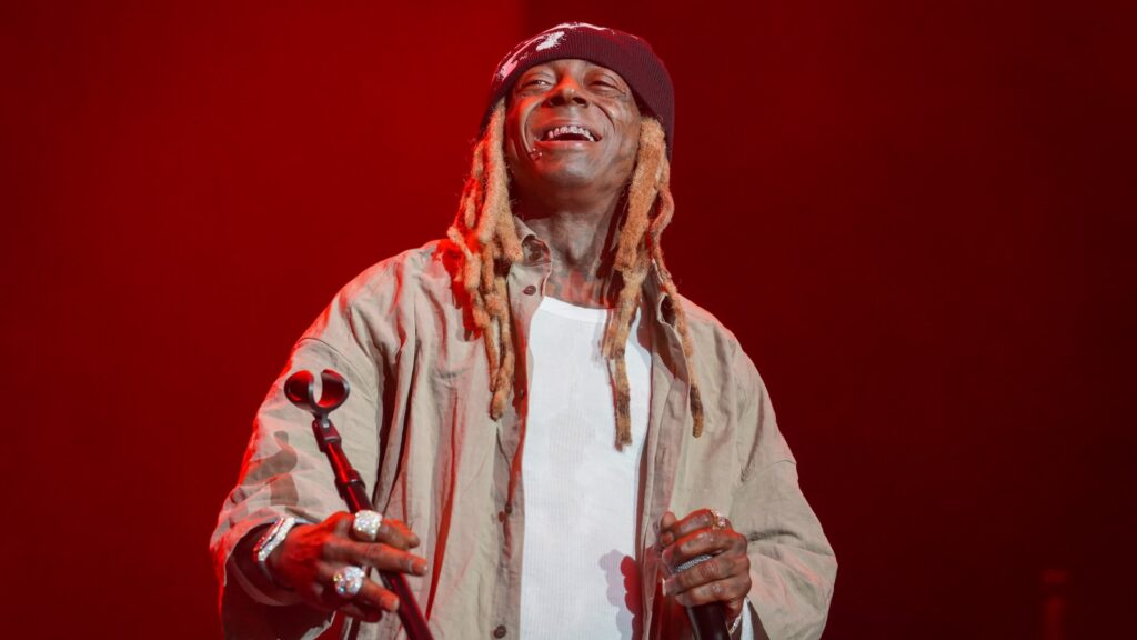 Lil Wayne Used AI to Recreate ‘Tha Carter’ Album Covers Lil Wayne performing on stage, wearing a beanie and casual outfit, smiling with a microphone in hand against a red-lit background.