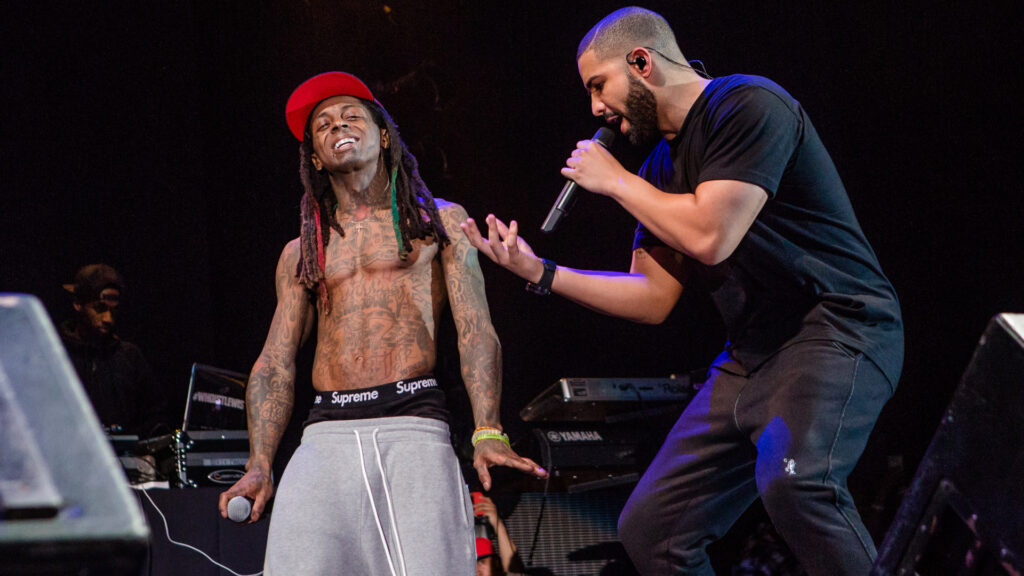 Lil Wayne and Drake performing on stage. Lil Wayne is shirtless, wearing a red cap and gray pants, while Drake holds a microphone.