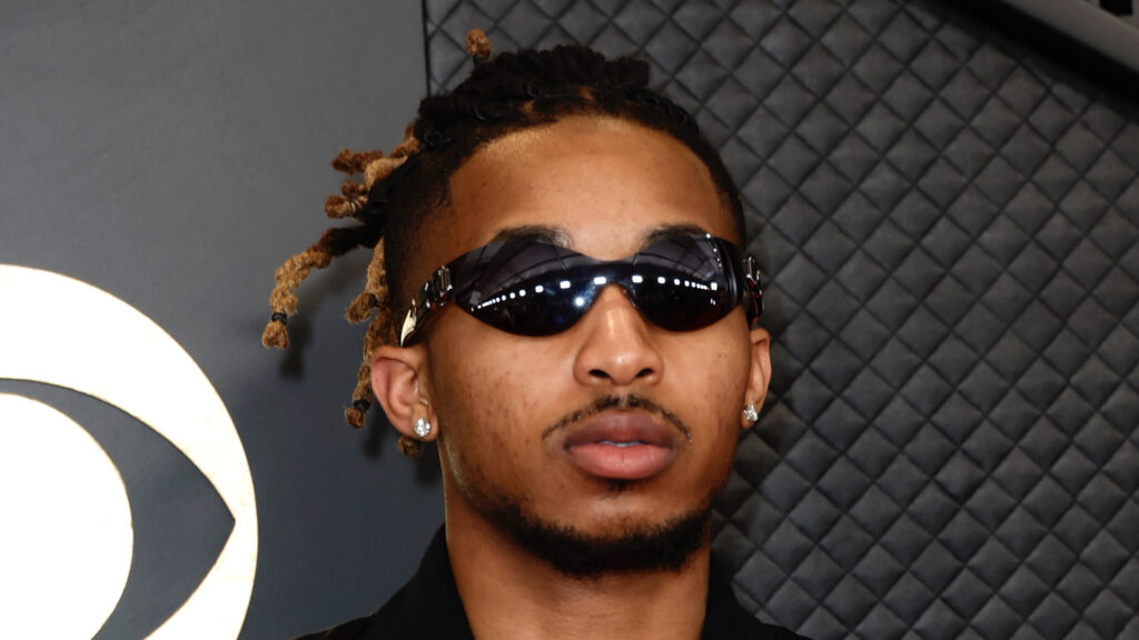 DDG wearing sunglasses and earrings, with braided hair, standing in front of a textured wall.