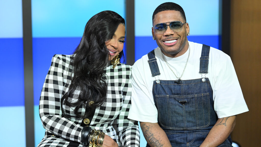 Ashanti in a checkered outfit and Nelly in overalls, both smiling and sitting together in a studio setting.