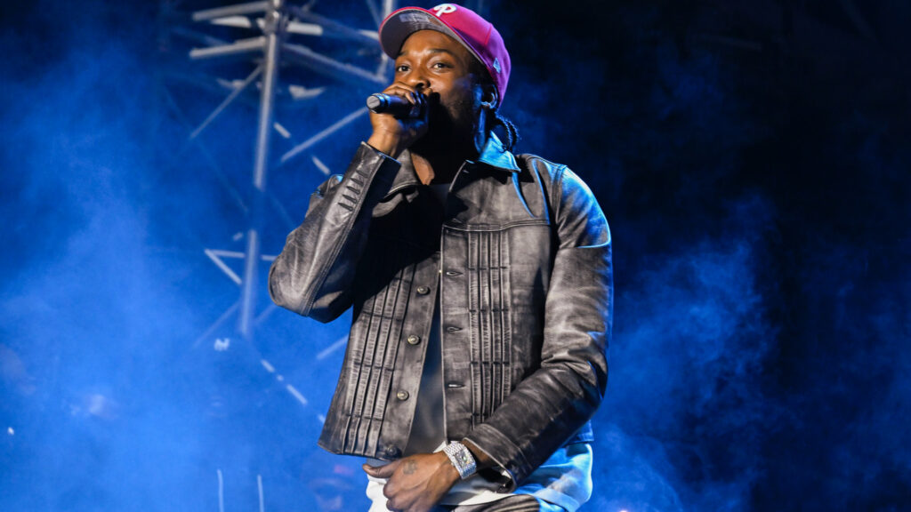 Meek Mill wearing a red cap and leather jacket is performing on stage with blue smoke in the background.