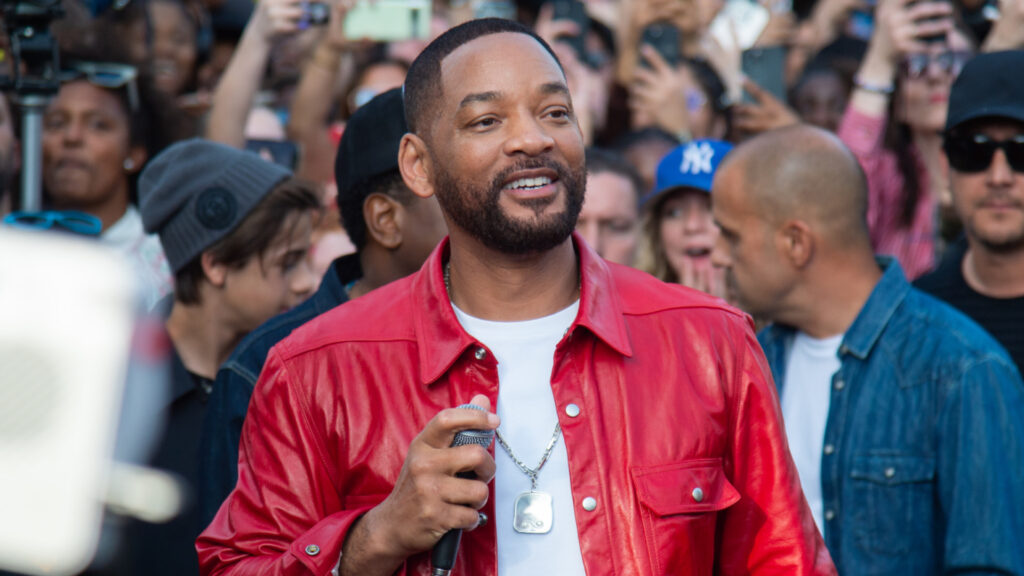 Will Smith in a red jacket, holding a microphone, surrounded by a crowd of fans.
