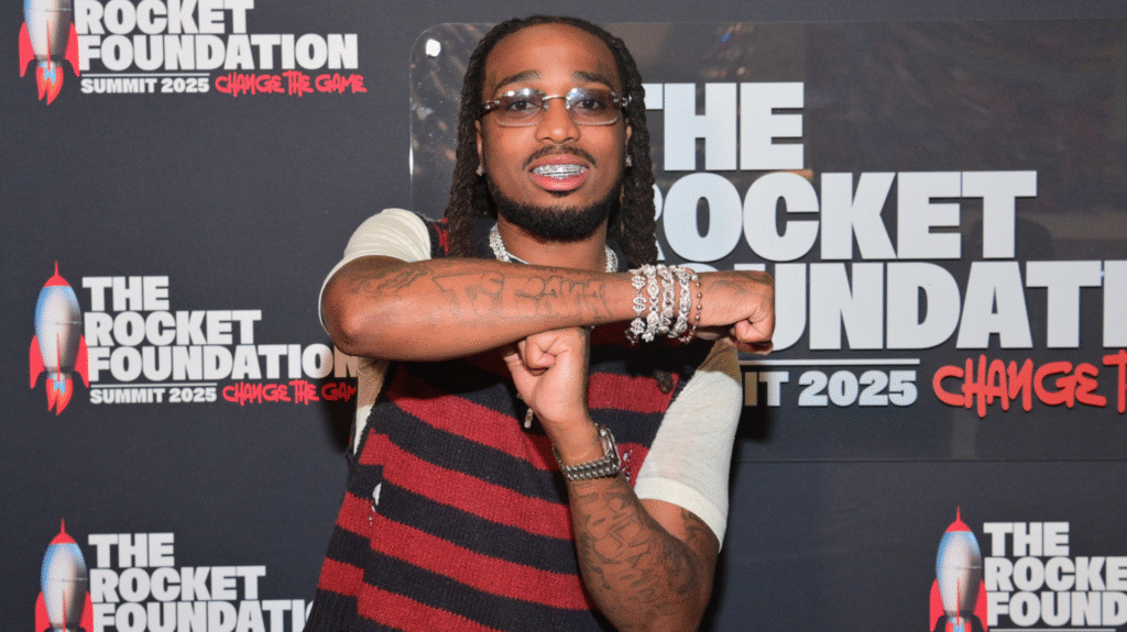 Quavo Hosts Second Annual Rocket Summit in Atlanta to Add… Quavo poses at The Rocket Foundation event, wearing glasses, a striped shirt, and jewelry, in front of a branded backdrop.