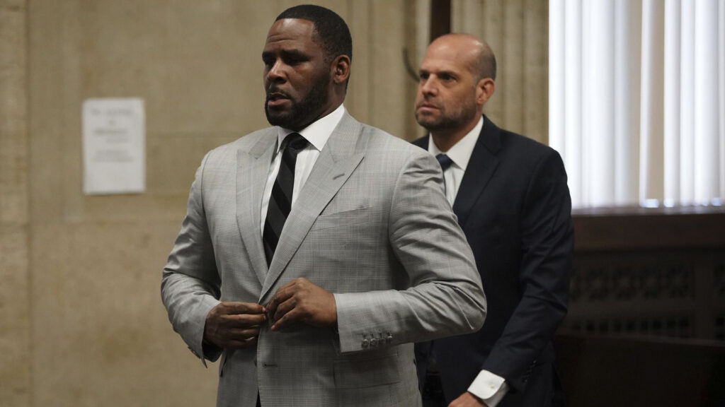 R. Kelly in a courtroom, wearing a light gray suit and black tie, accompanied by a man in a dark suit.