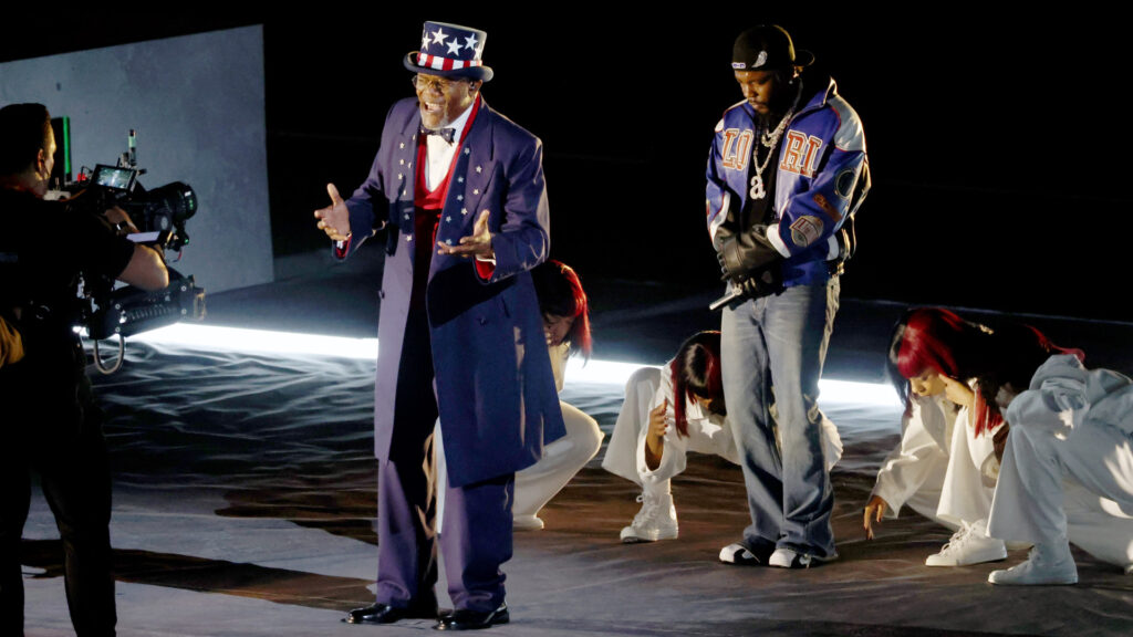 Samuel L Jackson in a patriotic outfit and another in casual clothing perform on stage with dancers and Kendrick Lamar. A camera operator films them.