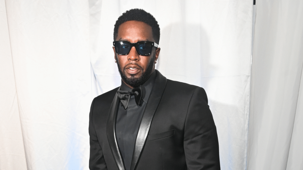 Sean "Diddy" Combs in a black tuxedo and sunglasses, standing against a white backdrop.