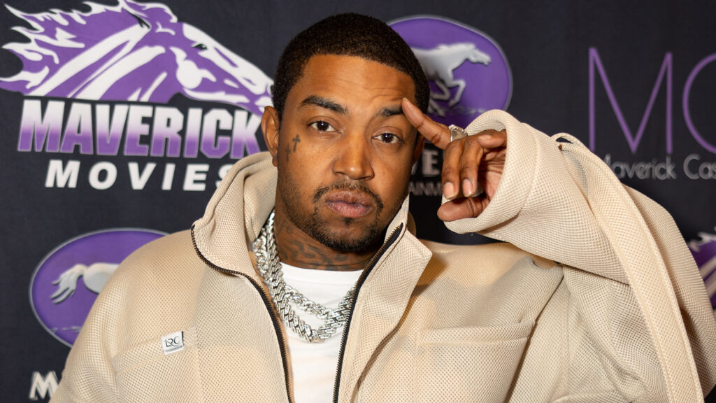 Lil Scrappy in a beige jacket poses in front of a backdrop with "Maverick Movies" text and logos.