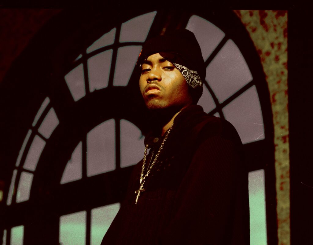 Nas wearing a dark beanie and bandana stands in front of an arched window, looking serious.