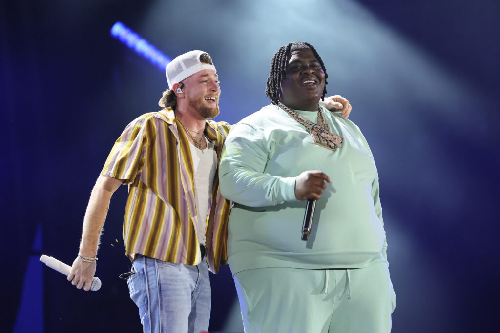 Bailey Zimmerman and BigXthaPlug on stage. Bailey is in a striped shirt and cap, BigXthaPlug is in a light green outfit. They are sharing a moment under bright stage lights.