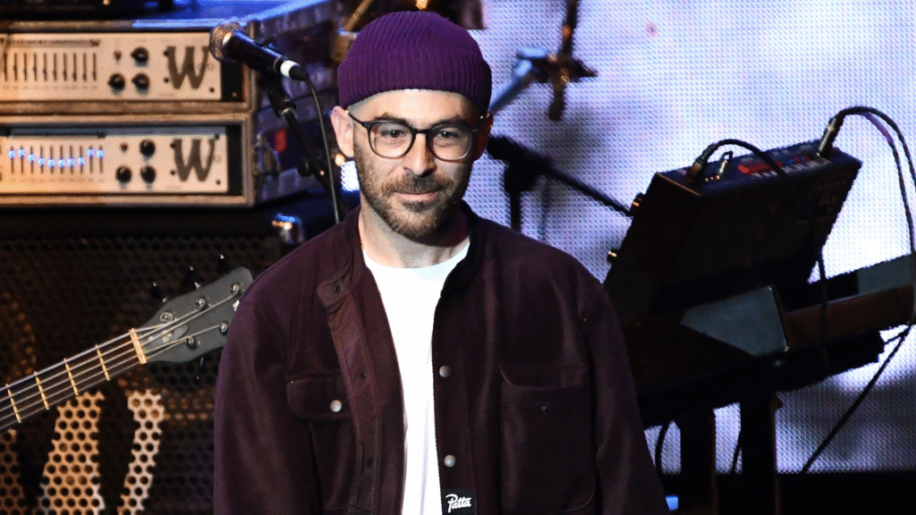 A man wearing glasses, a purple beanie, and a dark jacket stands on stage with musical equipment in the background.