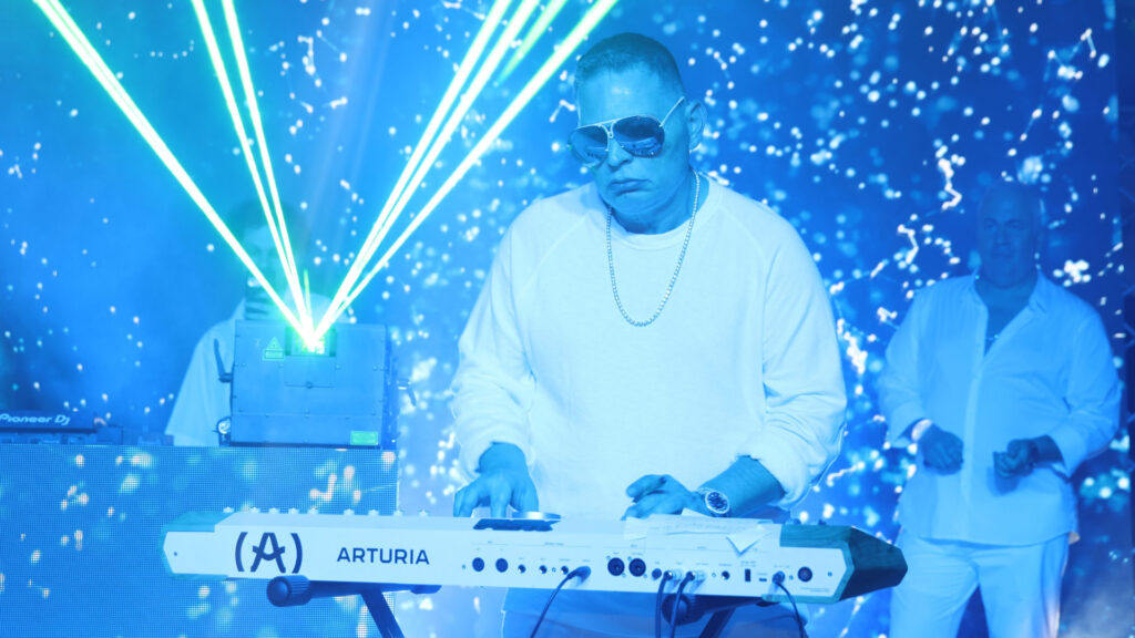 Scott Storch in sunglasses playing a keyboard with laser lights and a starry backdrop. Another person stands in the background.