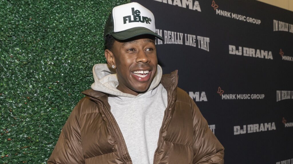 Tyler, the Creator, wearing a brown jacket and "le FLEUR" cap, smiles at a DJ Drama event backdrop.