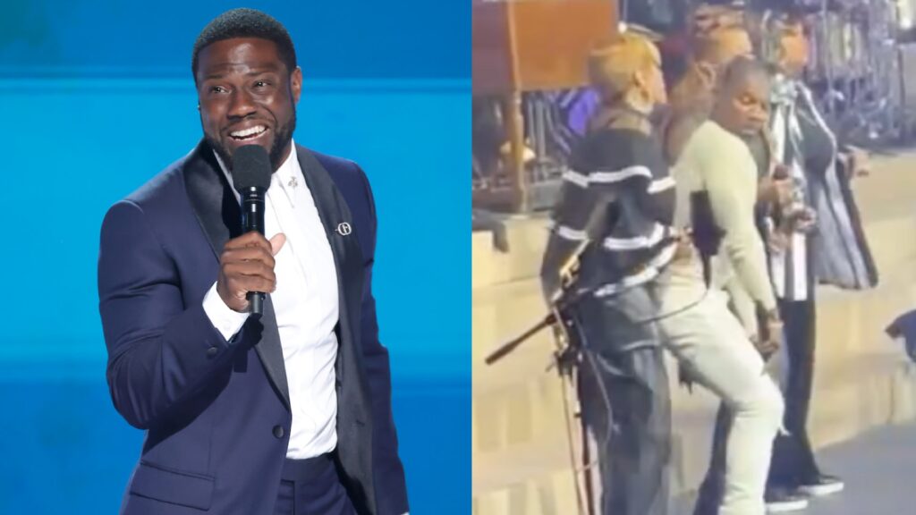 Kevin Hart, in a navy suit, holding a microphone and smiling on stage against a blue background.
