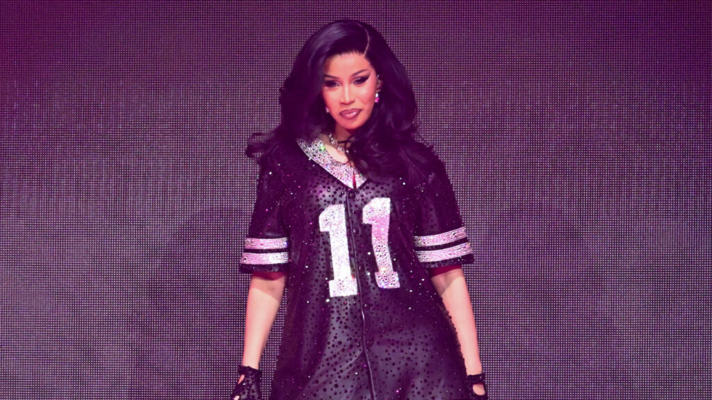 Cardi B performs in a sparkling black jersey with the number 11, set against a vibrant stage backdrop.