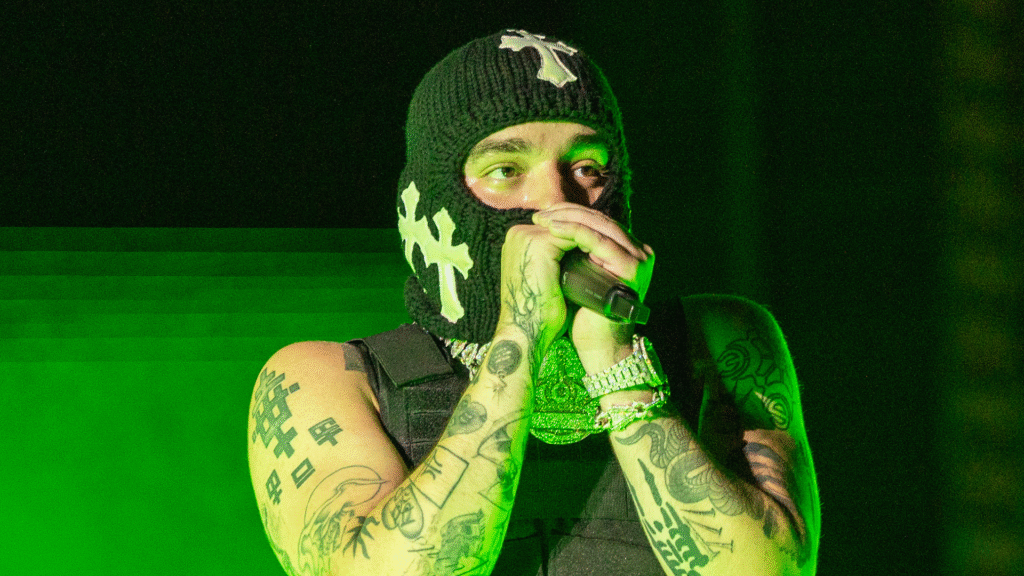 Yeat Surprises Nardwuar With a Handmade Mask A person wearing a black ski mask with white cross designs, performing on stage with tattoos on their arms, holding a microphone.