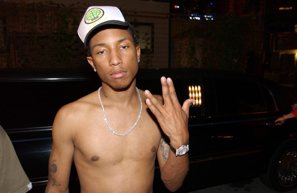 A shirtless Pharrell wearing a cap and chain necklace poses with a hand gesture in front of a black car.