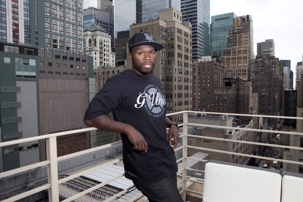 50 Cent stands on a balcony overlooking New York City, wearing a black G-Unit shirt and cap. Skyscrapers are in the background.