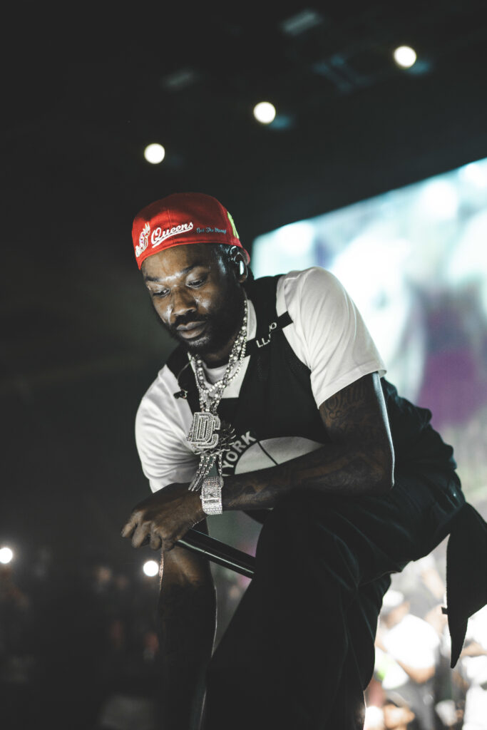 Meek Mill wearing a red cap and large chain, holding a microphone, with a blurred audience in the background.