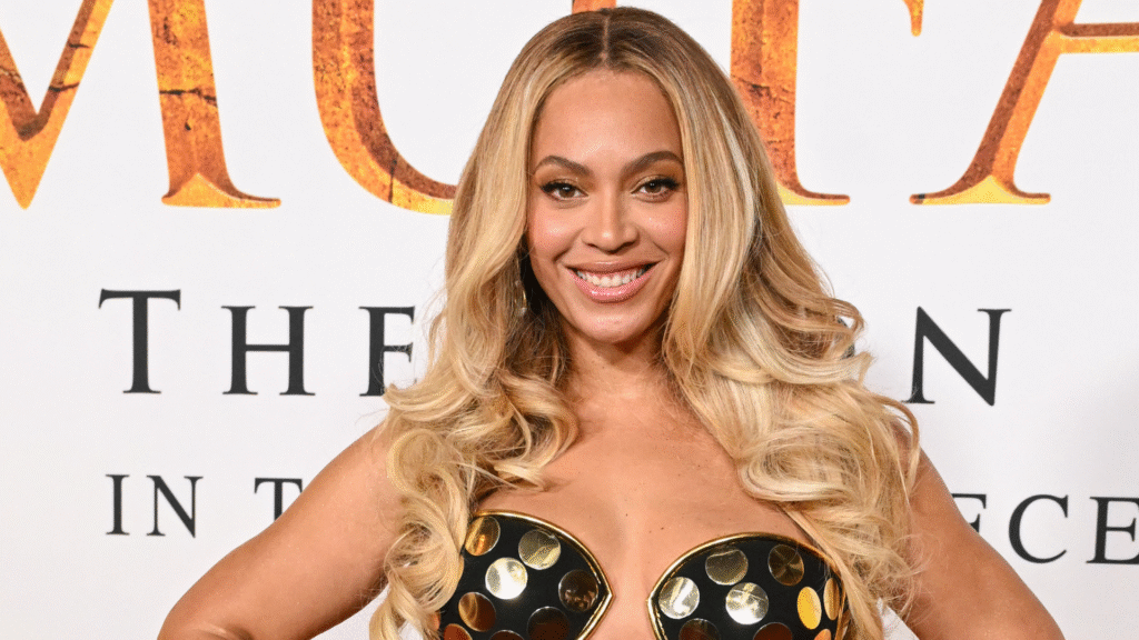 Beyoncé at an event, wearing a black and gold dress with long blonde hair, smiling at the camera.