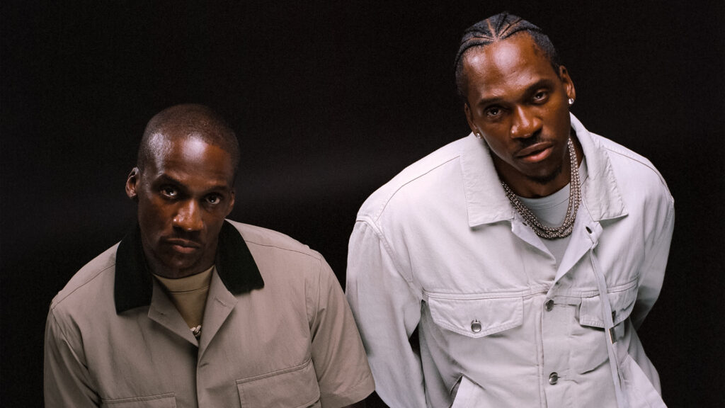 Clipse pose in stylish outfits against a dark background. Malice wears a tan jacket, Pusha T a white jacket with braids and a chain necklace.