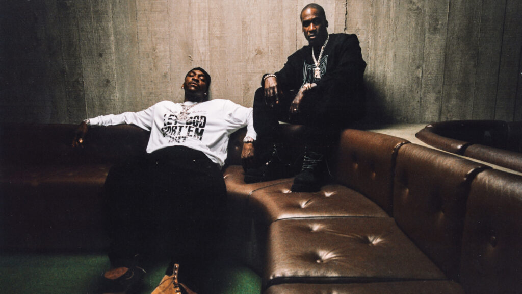 Clipse are sitting on a brown leather couch in a dimly lit room. Pusha T is reclining in a white shirt, Malice is sitting upright in black.
