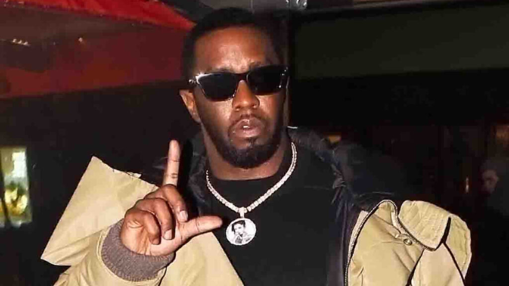 Sean "Diddy" Combs wearing sunglasses and a beige jacket, holding up a hand gesture.