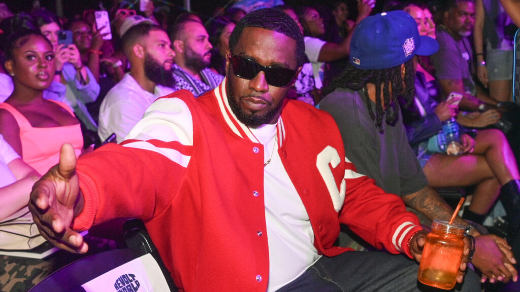 Sean "Diddy" Combs in a red jacket and sunglasses, sitting in a crowd at an event.