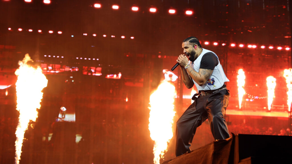 Drake performing energetically on stage, surrounded by flames, wearing a white vest and holding a microphone.