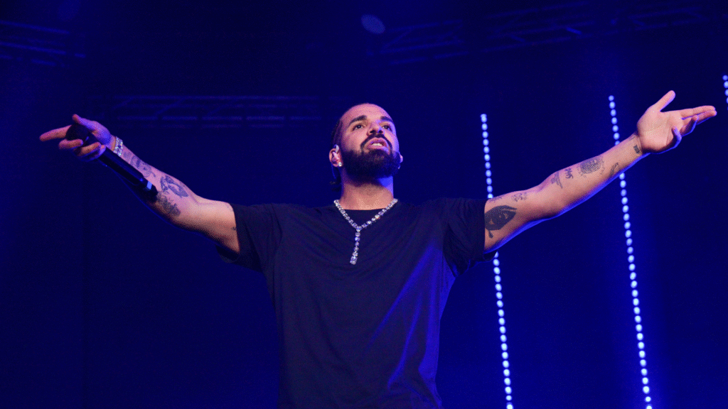 A man on stage with arms outstretched, wearing a black shirt and necklace, with tattoos on his arms, under blue stage lighting.