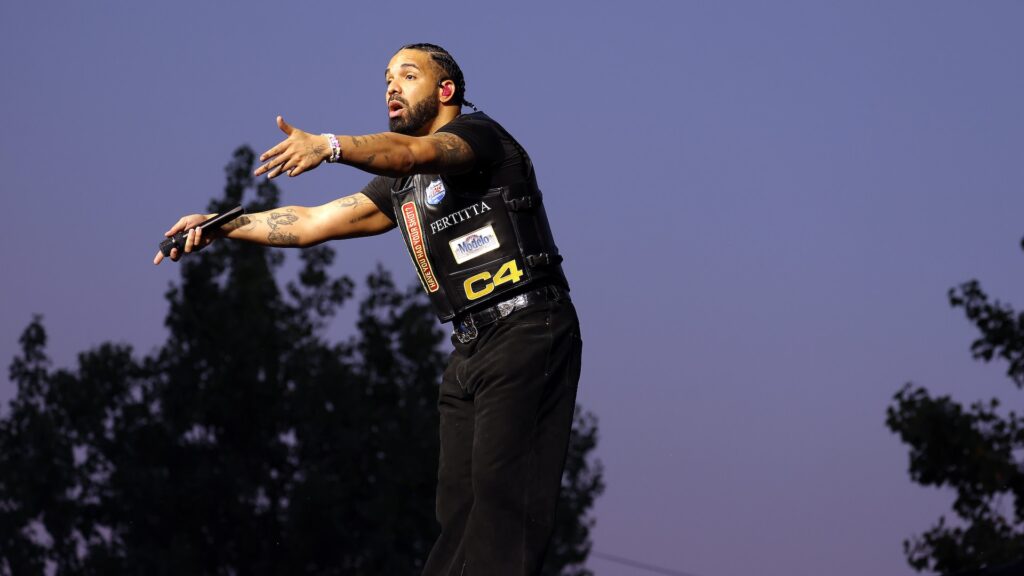 Drake performing on stage at Wireless Festival 2025, wearing a black vest with logos, gesturing passionately with a microphone in hand against a twilight sky.