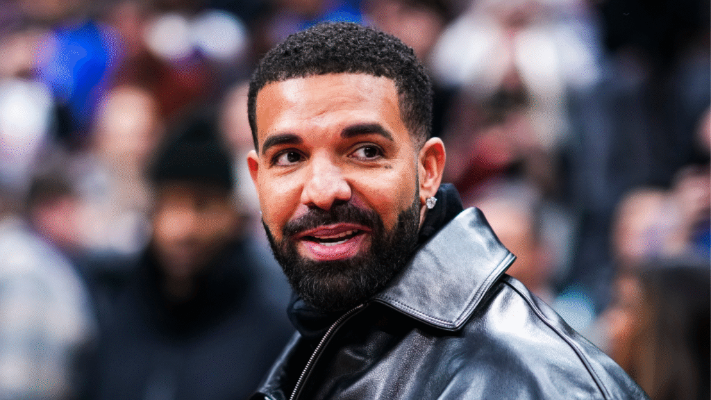 Drake in a black leather jacket, smiling, with a blurred crowd in the background.