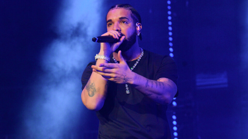 Drake performing on stage, holding a microphone, wearing a black shirt and jewelry, with a blue-lit background.