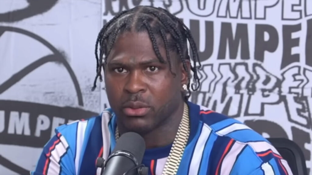 4xtra with braided hair and a striped shirt speaks into a microphone, with "NO JUMPER" text in the background.