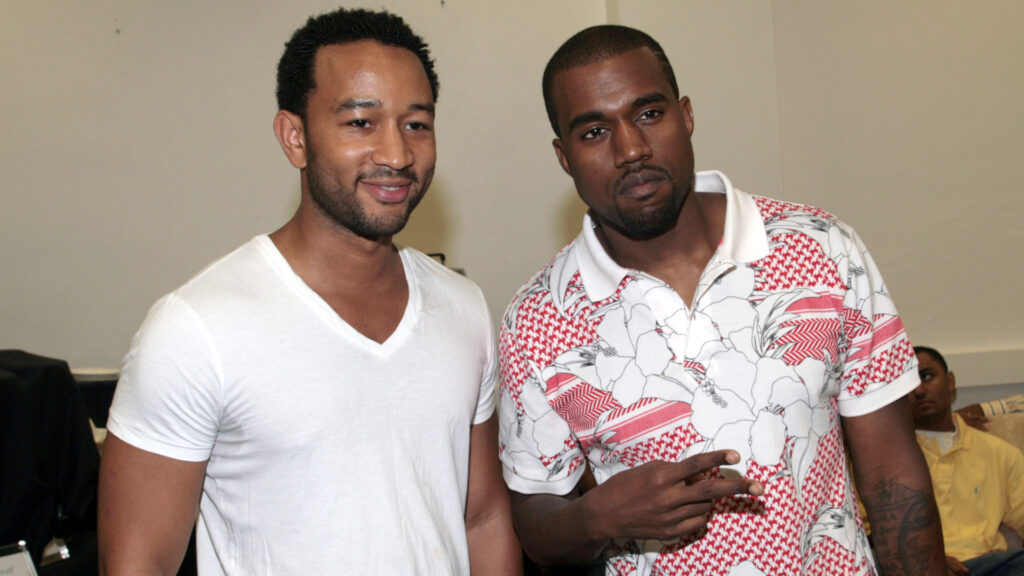 John Legend in a white t-shirt stands next to Kanye West, who is wearing a patterned polo shirt, both posing indoors.