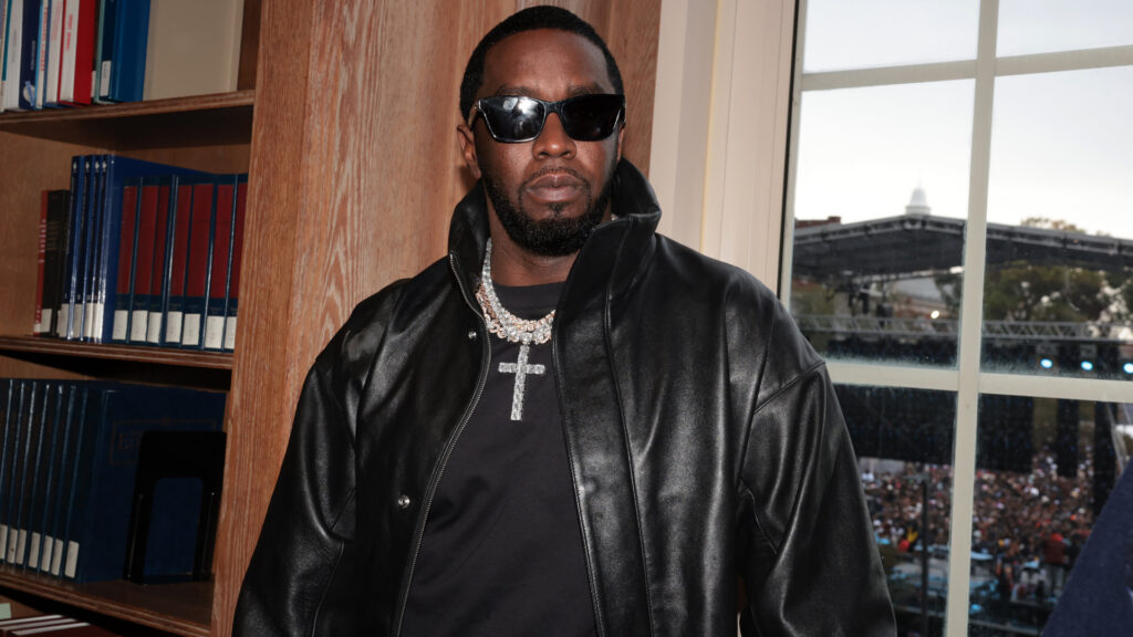 Sean Combs, wearing sunglasses and a black leather jacket, stands indoors with a crowd visible outside through the window.