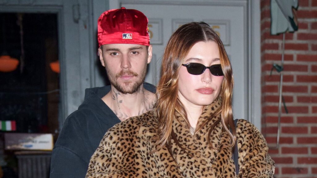 Justin Bieber in a red cap and Hailey Bieber in a leopard coat and sunglasses, walking together.