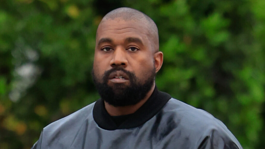 Ye (formerly Kanye West) with a beard and short hair wearing a dark top, standing outdoors with greenery in the background.