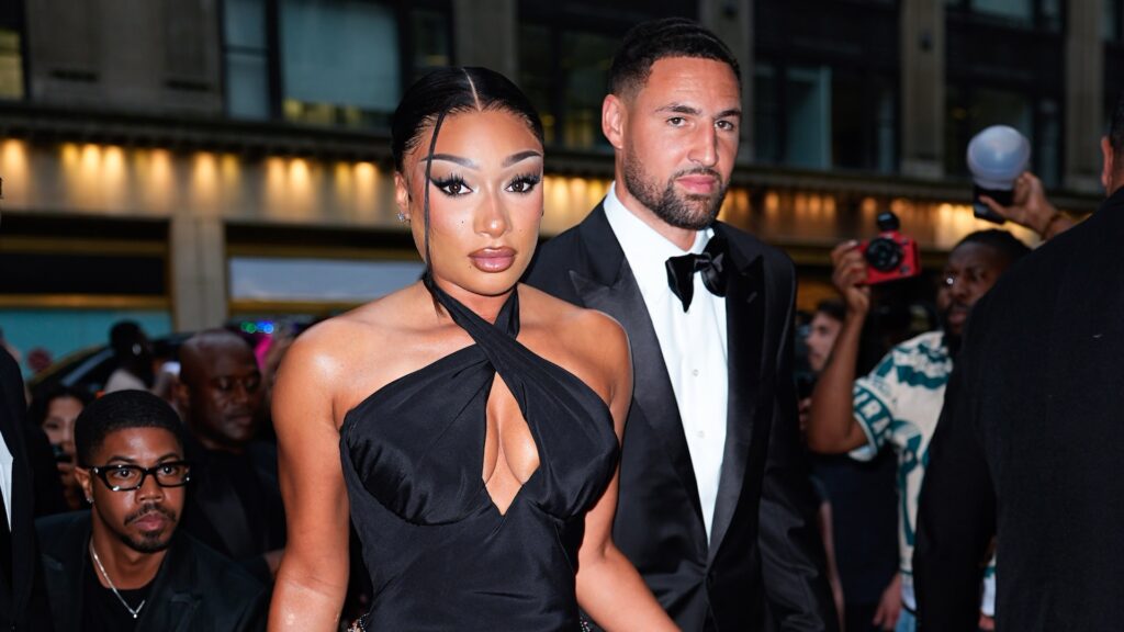 Megan Thee Stallion in a black dress and Klay Thompson in a tuxedo at a formal event, with onlookers and photographers in the background.