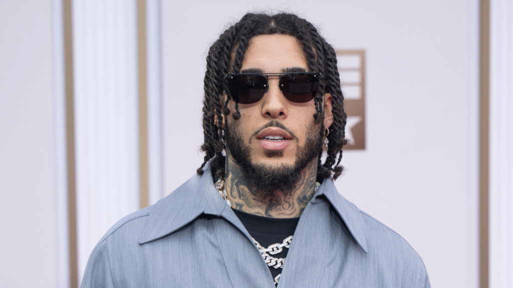 A man with sunglasses, wearing a light blue shirt and chain necklace, stands in front of a patterned background.