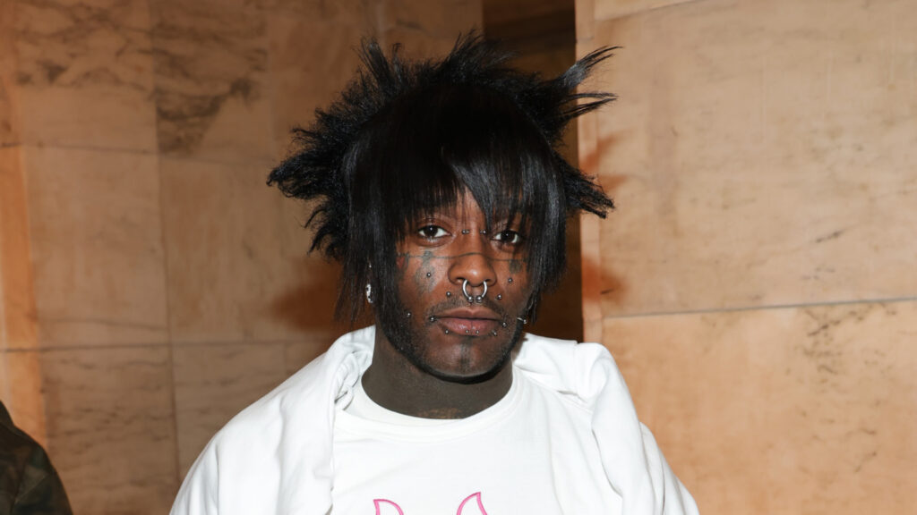 Lil Uzi Vert with spiky hair, facial piercings, and a white shirt, standing against a marble wall.