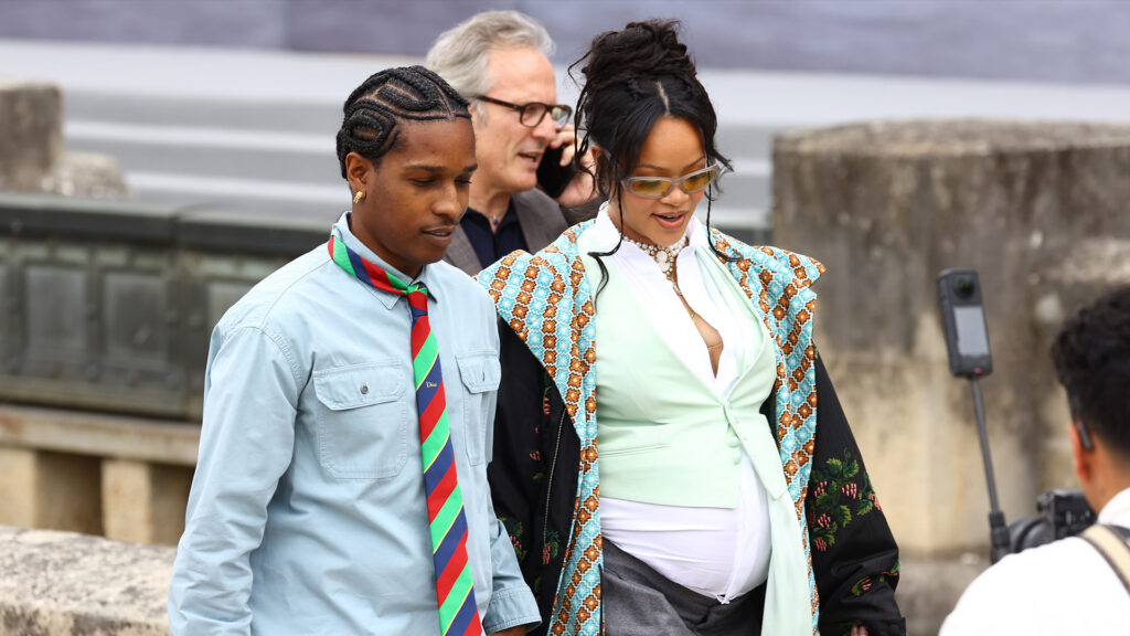 A$AP Rocky and Rihanna walking together. A$AP Rocky wears a blue shirt and striped tie, Rihanna wears a patterned coat and sunglasses.