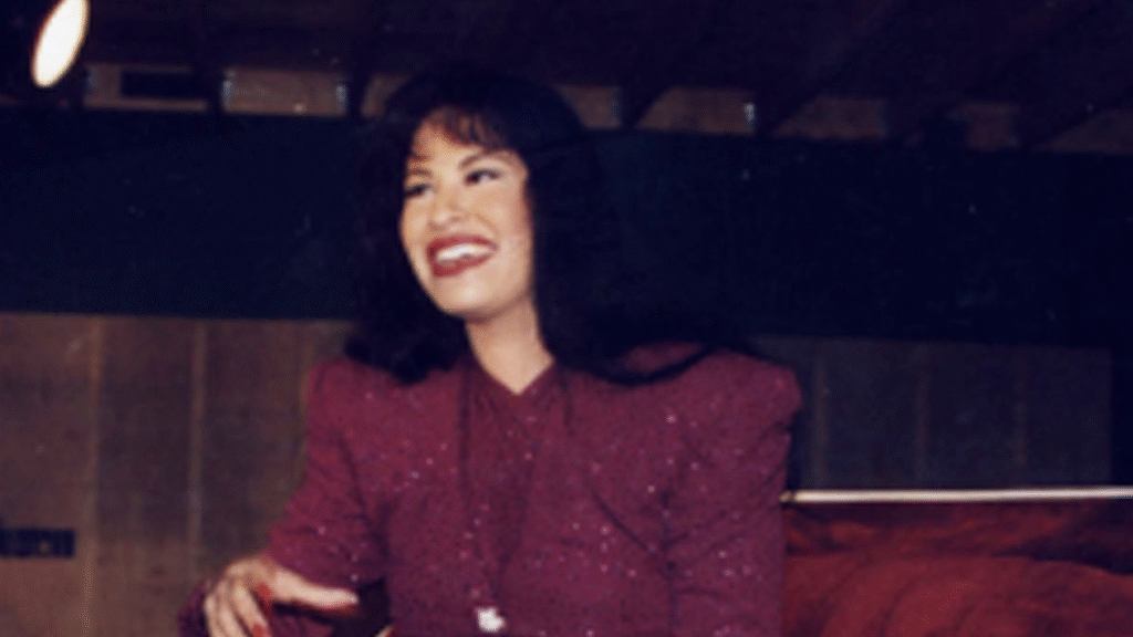 A woman with long dark hair and a sparkling burgundy outfit smiles broadly, seated indoors.