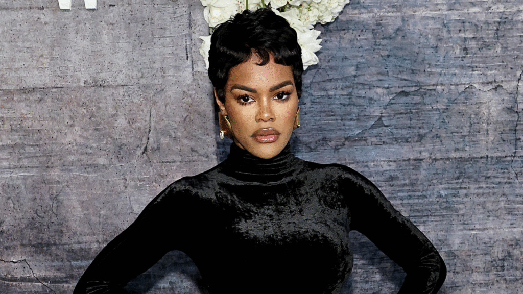 Teyana Taylor in a black velvet dress, posing confidently against a textured backdrop with white flowers above.