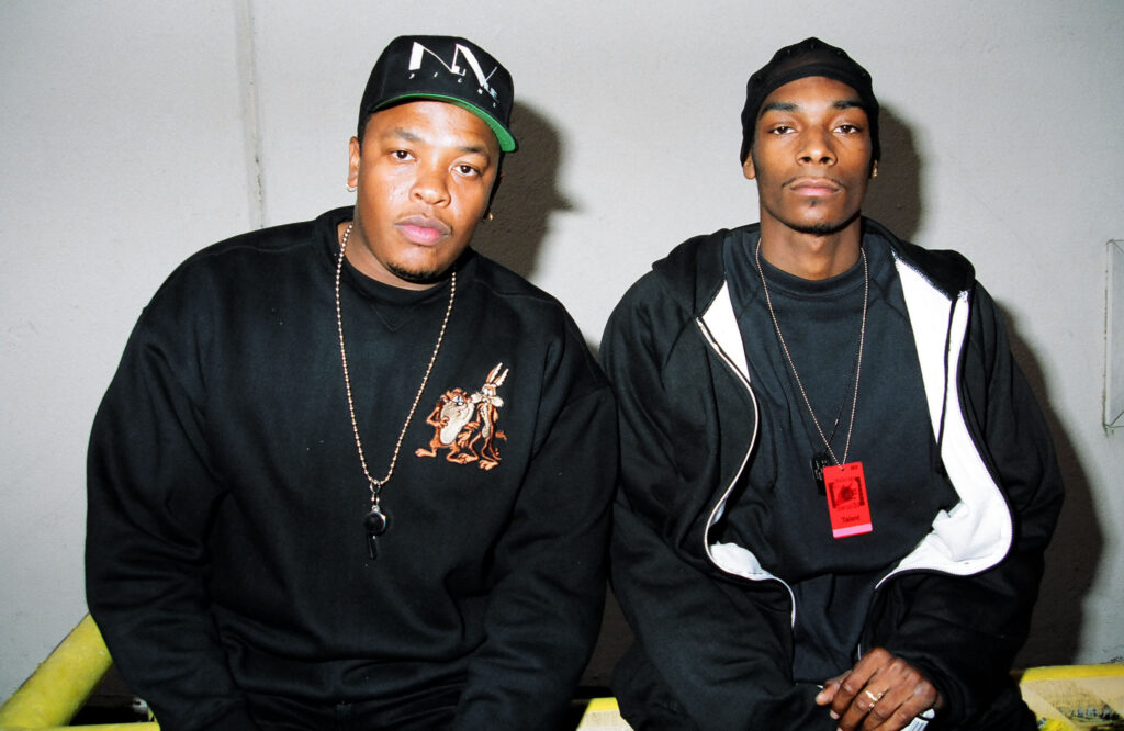 Dr. Dre and Snoop Dogg sitting together, both wearing black outfits. Dr. Dre has a cap, and Snoop Dogg wears a beanie.