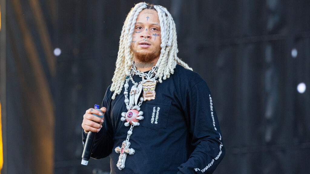 Rapper Trippie Redd performing on stage, wearing a black shirt and large chains, holding a microphone.