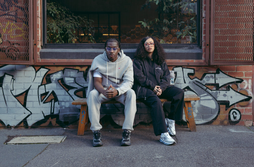 A.Girl and VV Ace sit on a bench in front of graffiti-covered wall. The person on the left wears a New Balance gray tracksuit, and the one on the right wears black.