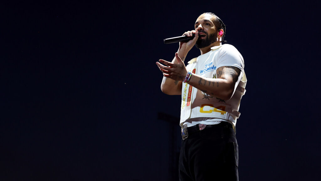 A man with tattoos and a beard performs on stage, holding a microphone, wearing a white t-shirt and black pants against a dark background.